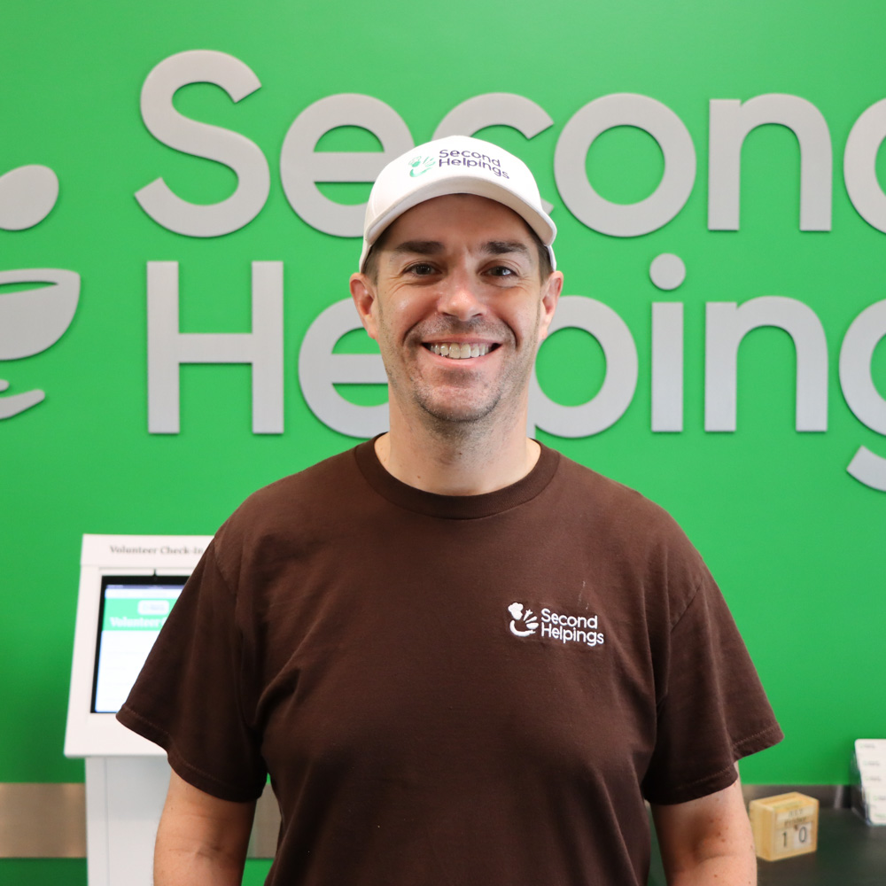Andrew, smiling, wearing a brown shirt and white hat with Second Helpings logos on them
