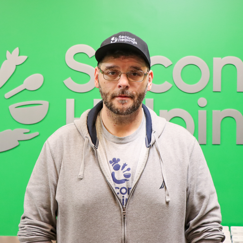 Zach with glasses wearing a grey zip up hoodie opened halfway to expose a Second Helpings logo on his shirt